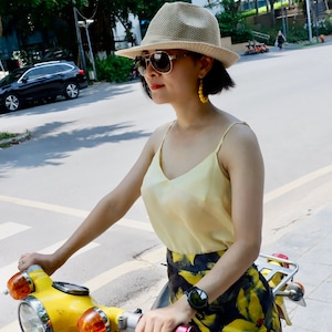Puede incluir: Una mujer con un top amarillo sin mangas, una falda floral negra y amarilla, un sombrero de paja y gafas de sol está conduciendo un ciclomotor amarillo. El ciclomotor tiene un logotipo de Honda en el lateral.