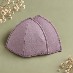 Puede incluir: Un par de copas de sujetador triangulares en un tono lavanda apagado. Cada copa presenta una superficie lisa cubierta de tela con una costura vertical central y un acabado de puntada overlock a juego a lo largo de los bordes curvos.