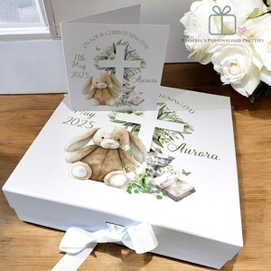 May include: White gift box with a white ribbon and a white card. The box has a design of a cross, a bunny, and greenery. The card says "On your christening day" and "11th May 2025 Aurora".