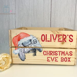 May include: Wooden Christmas Eve box featuring a festive design with a Santa hat on a video game controller. The box is personalized with the name "Oliver's" and the text "Christmas Eve Box" in red.