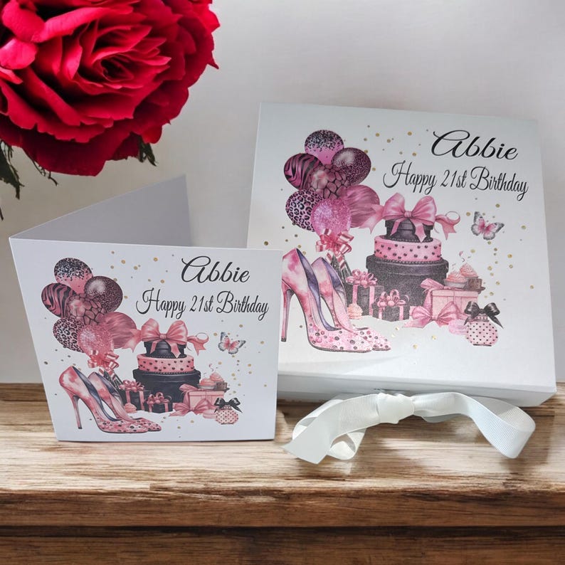 May include: A white gift box and card set with a birthday theme. The card and box feature illustrations of high heels, balloons, gifts, and a cake. The text "Abbie Happy 21st Birthday" is displayed on both items. A white ribbon is tied around the box.