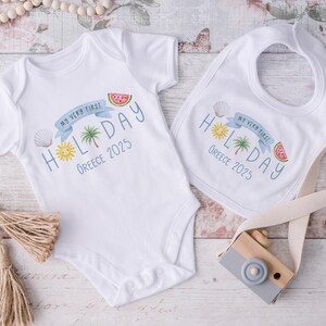 May include: White baby bodysuit and bib set. The bodysuit and bib feature a colorful design with the text "MY VERY FIRST HOLIDAY Greece 2025". The design includes a sun, palm tree, seashell, and watermelon slice.