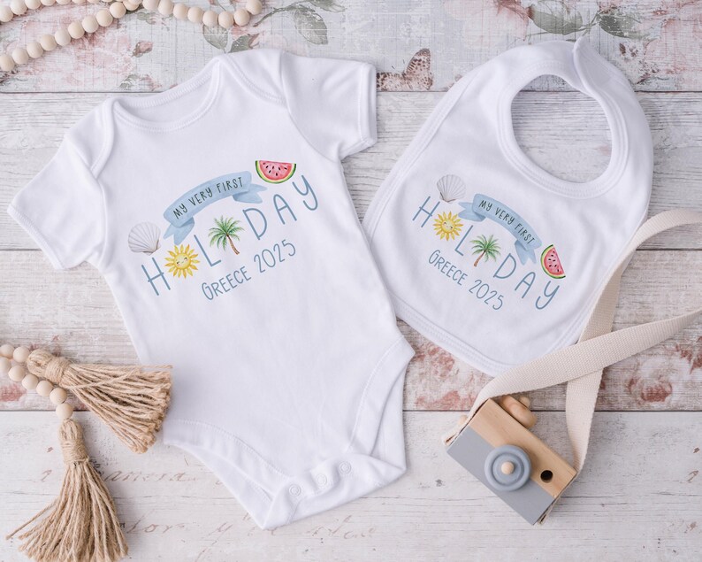 May include: White baby bodysuit and bib set. The bodysuit and bib feature a colorful design with the text "MY VERY FIRST HOLIDAY Greece 2025". The design includes a sun, palm tree, seashell, and watermelon slice.