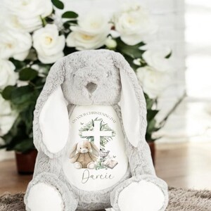 May include: A gray plush bunny with long ears. The bunny has a white patch on its chest with a cross, a small bunny, a chalice, a book, and the text "On your christening day Darcie".