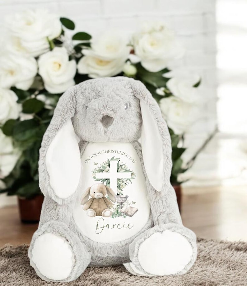 May include: A gray plush bunny with long ears. The bunny has a white patch on its chest with a cross, a small bunny, a chalice, a book, and the text "On your christening day Darcie".