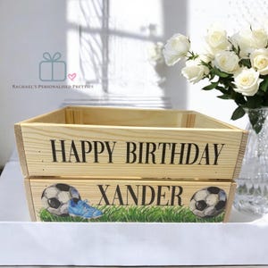 May include: A wooden crate with the text "HAPPY BIRTHDAY XANDER". The crate is decorated with footballs, a blue boot, and green grass. A bouquet of white roses is in a vase in the background.