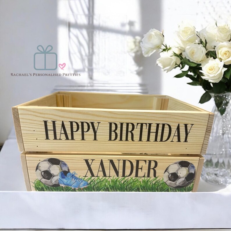 May include: A wooden crate with the text "HAPPY BIRTHDAY XANDER". The crate is decorated with footballs, a blue boot, and green grass. A bouquet of white roses is in a vase in the background.