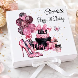 May include: A white gift box with a bow, decorated with pink and black illustrations. The design includes high heels, balloons, a cake, and the text "Charlotte Happy 16th Birthday". Gold confetti dots the background.
