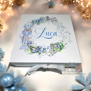 May include: White gift box with a watercolour design. It features a blue nutcracker, a wreath of blue berries and greenery, and the name "Luca" in blue script. A silver ribbon is tied around the box.