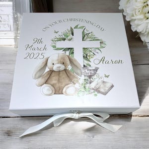 May include: White gift box with a light brown bunny, a cross, and flowers. The box has the text "On your christening day 9th March 2025 Aaron".