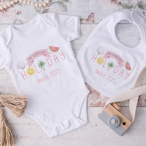 May include: White baby bodysuit and bib set with a pink banner that reads "My Very First Holiday" with a sun, palm tree, seashell, and watermelon graphic. The text "Greece 2025" is below the banner. A wooden toy camera is next to the set.