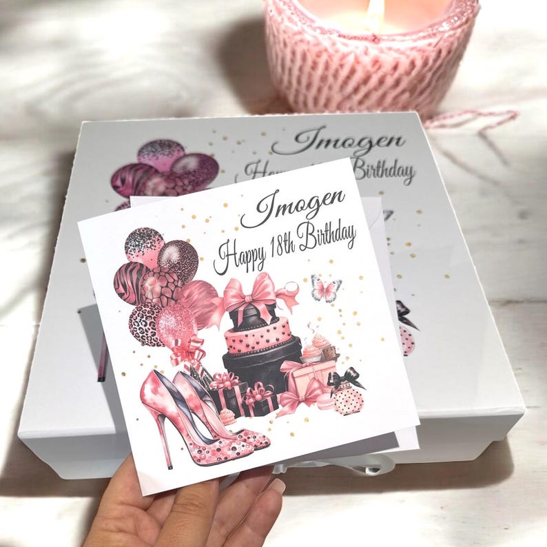 May include: A white gift box with a greeting card that reads "Imogen Happy 18th Birthday." The card features illustrations of pink high heels, a cake, gifts, and balloons. A lit candle sits in the background.