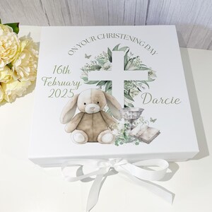 May include: White gift box with a green cross, flowers, and a bunny rabbit. The box has the text "On Your Christening Day" and "Darcie" on it. The date "16th February 2025" is also printed on the box.