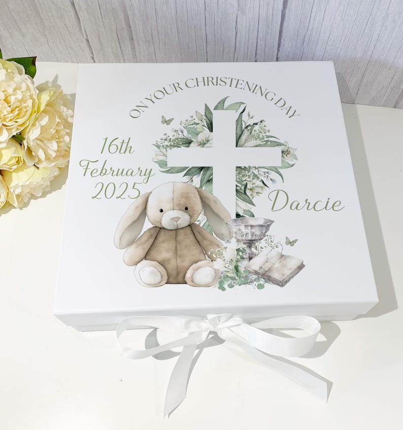 May include: White gift box with a green cross, flowers, and a bunny rabbit. The box has the text "On Your Christening Day" and "Darcie" on it. The date "16th February 2025" is also printed on the box.