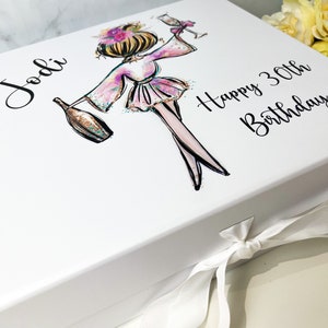 K&ouml;nnte beinhalten: Wei&szlig;e Geschenkbox mit wei&szlig;em Band. Die Box zeigt eine bunte Illustration einer Frau, die ein Glas Champagner h&auml;lt. Der Text "Jodi Happy 30th Birthday" ist auf der Box aufgedruckt.