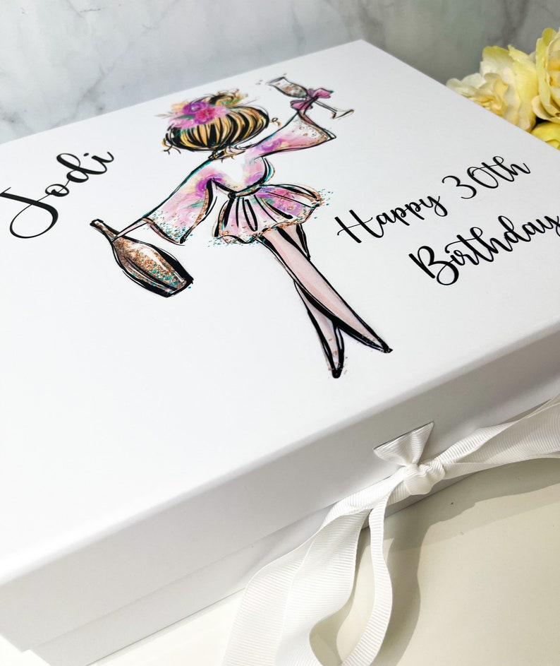 K&ouml;nnte beinhalten: Wei&szlig;e Geschenkbox mit wei&szlig;em Band. Die Box zeigt eine bunte Illustration einer Frau, die ein Glas Champagner h&auml;lt. Der Text "Jodi Happy 30th Birthday" ist auf der Box aufgedruckt.