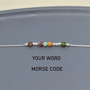 May include: A delicate silver chain bracelet featuring a series of colorful gemstone beads. The beads are in shades of brown, pink, green, and orange. The text "YOUR WORD MORSE CODE" is displayed below the bracelet.