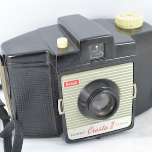 Puede incluir: Cámara Kodak Brownie Cresta II vintage. La cámara negra tiene una cara plateada y blanca con el logotipo de Kodak. Tiene una lente negra y una correa negra. La cámara mide aproximadamente 13 cm de largo.