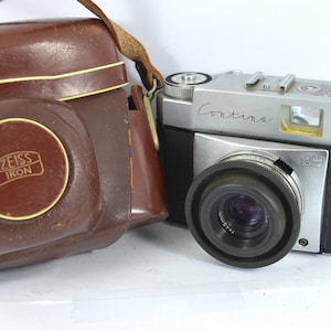 May include: A vintage Zeiss Ikon Contina camera with a brown leather case. The camera is silver and black with a black lens and the word "Contina" written on the top. The leather case has the Zeiss Ikon logo.