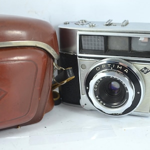 Puede incluir: Una cámara Agfa Optima vintage con cuerpo plateado y negro, y una funda de cuero marrón. La lente de la cámara está etiquetada como "Optima Color Apotar". La funda tiene un ribete plateado y un logotipo.