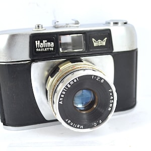 May include: A vintage Halina Paulette camera with a silver top and black leatherette body. The lens is marked Anastigmat 1:2.8 F=45mm. The camera has a viewfinder and a crown emblem. A classic film camera.