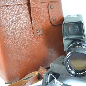 May include: A vintage Canon Zoom 8mm movie camera with a brown leather case. The camera is gray and black with a zoom lens. The case is rectangular and has a handle and metal hardware.