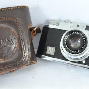 Op de afbeelding: Een vintage Halina Anastigmat camera met een zwarte behuizing en zilveren accenten, naast een bruine leren etui. De cameralens is gemarkeerd met "Halina Anastigmat f-3.5 45mm". De leren etui heeft het Halina logo.