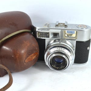 May include: A silver and black 35mm film camera with a leather case. The camera has a lens labeled "Voigtlander Color Skopar 1:2.8 50mm".