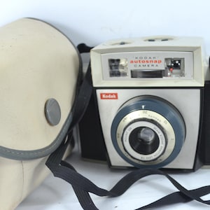 以下が含まれることがあります： クリーム色のボディと青いレンズリングを備えた、ヴィンテージのKodak Autosnapカメラ。 カメラには黒いストラップが付いており、クリーム色のケースが付属しています。 Kodakのロゴが見えます。