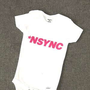 Puede incluir: Un body blanco para bebé con una estrella rosa y el texto "*NSYNC" impreso en la parte delantera.