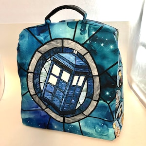May include: A blue and white tote bag with a stained glass design featuring a blue police box. The bag has a black handle and a blue and white starry sky background.