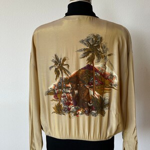 May include: A beige silk bomber jacket with a black ribbed collar and cuffs. The back of the jacket features a tropical print of palm trees, a volcano, and two figures.