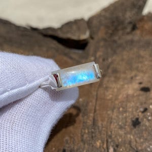 May include: A rectangular, iridescent moonstone pendant with silver-colored metal accents. The stone displays a play of blue and white light. The pendant is attached to a white cord, set against a natural, textured background.