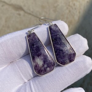 May include: A pair of earrings featuring rectangular, faceted purple gemstones set in silver-colored metal. The earrings have a hook closure and are displayed against a white background.