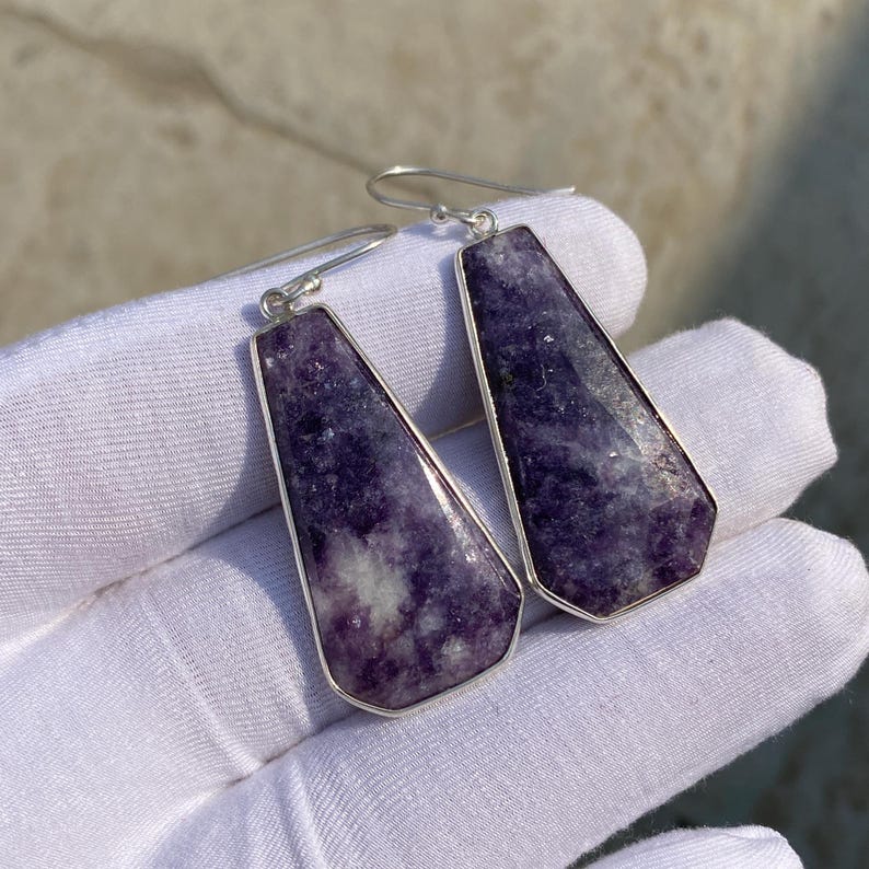May include: A pair of earrings featuring rectangular, faceted purple gemstones set in silver-colored metal. The earrings have a hook closure and are displayed against a white background.