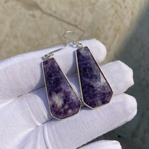 May include: A pair of earrings featuring a rectangular, trapezoid-shaped purple stone with a silver-colored metal border. The earrings have a hook-style closure. The stone has a mottled appearance with white and darker purple areas.