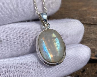 May include: Oval moonstone pendant necklace with a silver chain. The moonstone has a pearly white base with iridescent blue and yellow flashes. The pendant is set in a silver bezel. The necklace is displayed on a white surface.