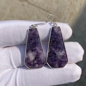 May include: A pair of earrings featuring a trapezoid-shaped, purple stone with white inclusions, set in a silver-colored metal frame. The earrings have a hook closure.