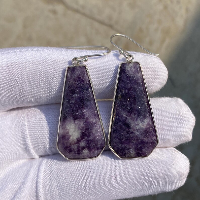 May include: A pair of earrings featuring a trapezoid-shaped, purple stone with white inclusions, set in a silver-colored metal frame. The earrings have a hook closure.