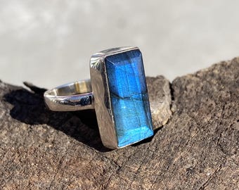 May include: A rectangular silver ring featuring a large, vibrant blue gemstone. The gemstone has a polished, reflective surface, showcasing shades of blue. The ring is set against a natural, textured wooden surface.
