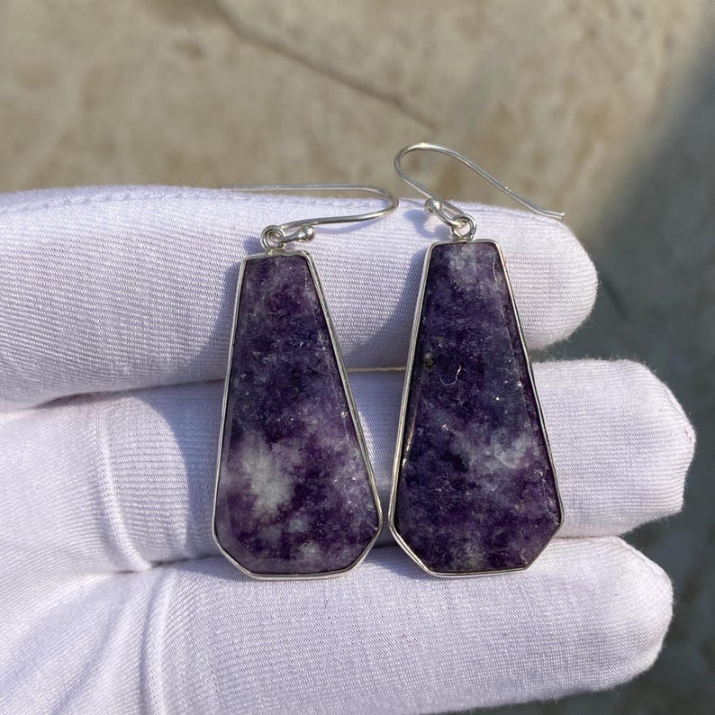 May include: A pair of earrings featuring a rectangular, trapezoid-shaped purple stone with white marbling, set in a silver-colored metal frame. The earrings have a hook closure. The background is a white surface.