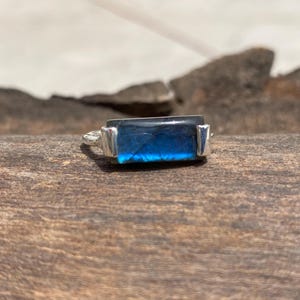 May include: A silver ring featuring a rectangular, iridescent blue gemstone. The gemstone has a dark blue base with lighter blue and teal flashes. The ring's band and setting are silver-colored.