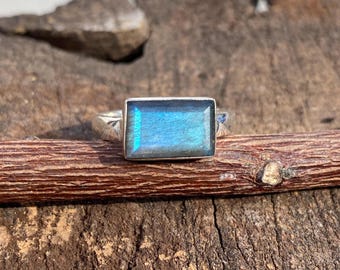 May include: A silver ring featuring a rectangular, iridescent blue gemstone. The ring is set on a wooden surface, showcasing the gemstone's color variations. The ring's band is simple and elegant, highlighting the stone's beauty.