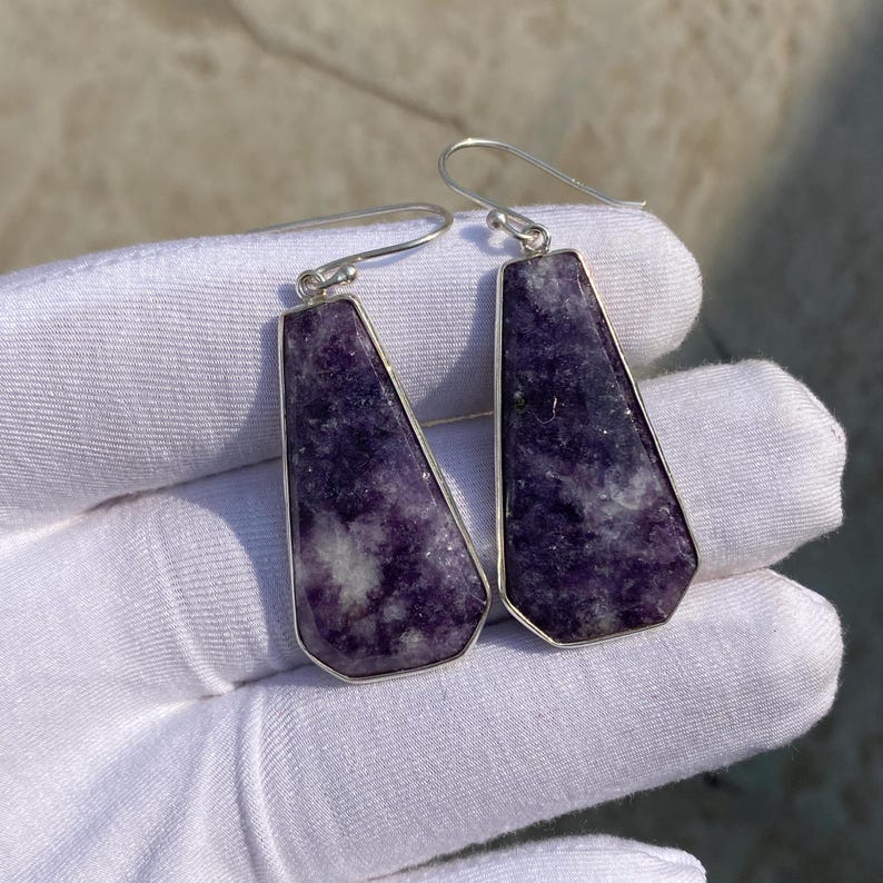 May include: A pair of earrings with a geometric shape. The earrings feature a vibrant purple stone with white and gray marbling, set in a silver-colored metal frame. The earrings have a hook closure.