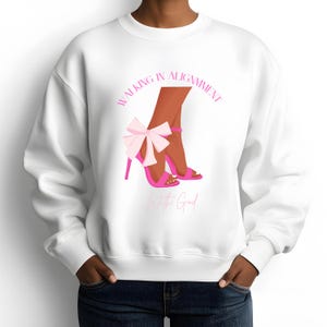 May include: White sweatshirt featuring a graphic of brown legs wearing pink high heels with a bow. The text "WALKING IN ALIGNMENT With the God" is printed in pink above and below the graphic. The sweatshirt has a crew neck and long sleeves.