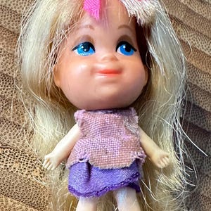 May include: A small doll with blonde hair, blue eyes, and a pink bow. She is wearing a purple dress with a pink vest.