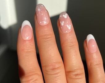 Op de afbeelding: Een hand met ovale nagels, sommige met een French manicure en witte punten. Andere nagels hebben een lichtroze basis met witte Mickey Mouse-hoofdontwerpen en sterrenpatronen. De nagels zijn netjes verzorgd en gepolijst.