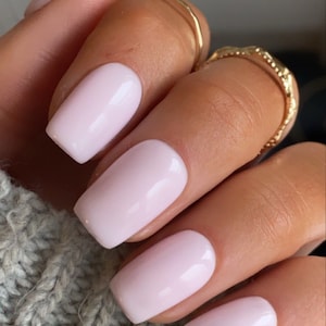 May include: Close-up of a hand with light pink, square-shaped acrylic nails. The nails are glossy and well-manicured. Two gold rings adorn the finger. The background is a gray knitted fabric.