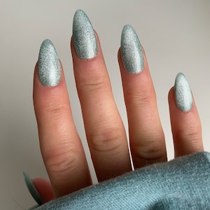 May include: Close-up of a hand with long, oval-shaped, light teal-colored fingernails. The nails have a shimmering, glittery finish. The hand is wearing a teal-colored long-sleeved shirt.