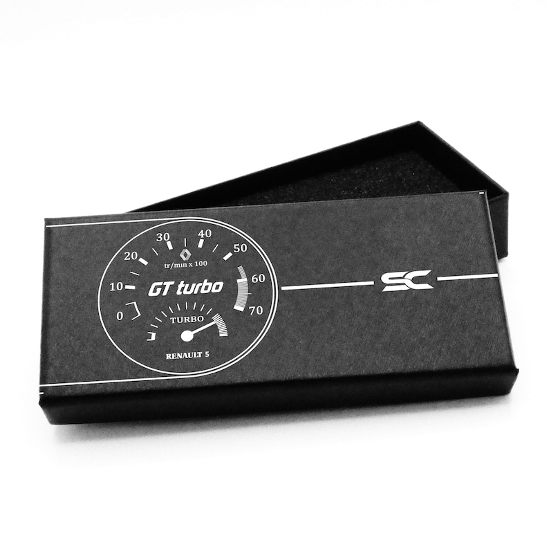 Puede incluir: Una caja rectangular negra con un gr&aacute;fico blanco de un medidor turbo de coche. El medidor indica "GT turbo" y "Renault 5". La caja est&aacute; abierta, revelando un interior negro. El logotipo "SC" est&aacute; en el lado derecho.